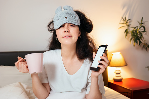 Portrait of sleeping cute young woman wearing sleep mask holding cup and phone in hands while sitting in bed. Concept of being late for work and morning awakening.