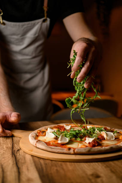istock Man in apron sprinkles arugula on pizza on a wooden stand before serving 2057476937