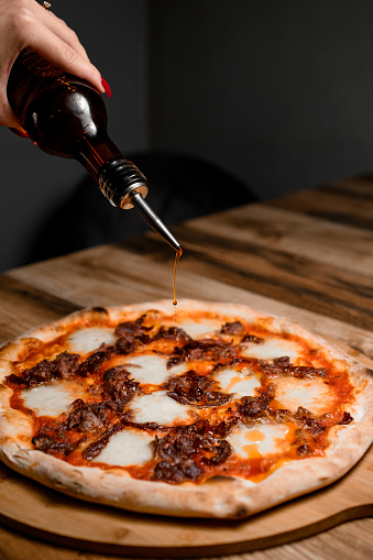 Female hand pours sauce on pizza with delicious cheese toppings on a round wooden stand on the table