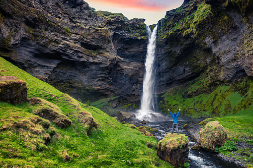 Colorful morning view of Kvernufoss waterfall with man standing on the rock. Majestic sunrise in south Iceland, Europe. Artistic style post processed photo.