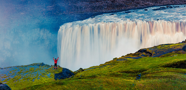 Tourist standing on the shore of falling water of the most powerful waterfall in Europe - Dettifoss with raised hands. Colorful summer sunset in Jokulsargljufur National Park, Iceland. White nights view.