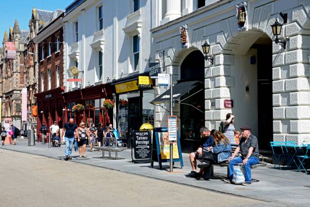 People sitting on a bench by a bus stop with shops and shoppers along Queen Street in the city centre, Exeter, Devon, UK, Europe.