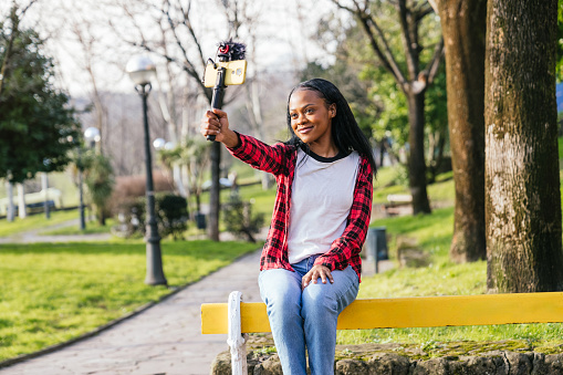 Smiling Afro-American films her walk in the park, selfie stick in hand.