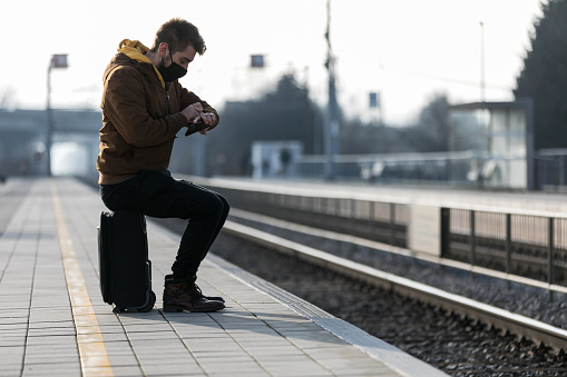 Young man sitting on black suitcase looking at his watch at railway station travel concept.