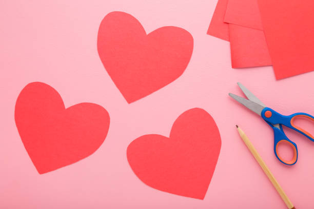 istock Red heart shapes, scissors, pencil and application paper on light pink table background. Pastel color. Closeup. Making decoration elements. Top down view. 2057600491