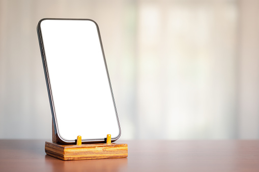 A smartphone with a blank white screen on a wooden stand against a blurred background, bathed in soft sunlight, exemplifies the seamless integration of technology and communication in our daily lives