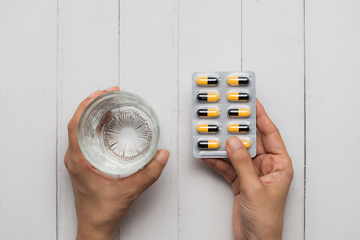 Top view of a hand holding a glass of water and a blister pack of pills against a white wooden background, illustrating health care routine or medication regimen