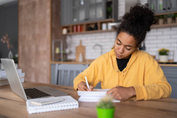 istock An african-american young woman is using laptop computer for remote work or studying online, she takes notes watching webinars or classes at kitchen table at home 2057671739