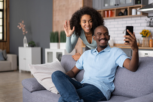 Remote Communication. African American man and woman waving palms at a mobile phone, sitting on the sofa in kitchen room interior. Cheerful black couple having video call via cellphone, staying at home