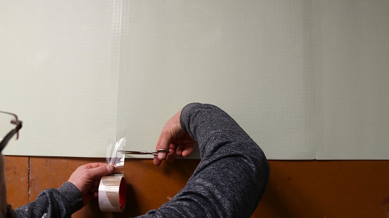 a man uses scissors to cut off a strip of tape glued at the junction of two polyester sheets of soft insulation on the floor, preparing and fixing the underlay before laying the laminate