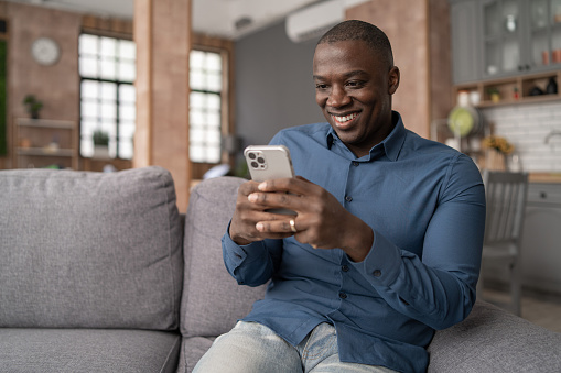 Excited Black African American Man Using Smartphone for mobile shopping while Sitting on the Sofa in Living Room. Happy Man buying things, shopping online, using mobile app
