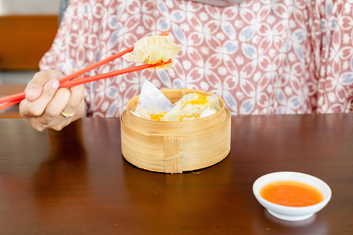 A woman is eating dim sum dumplings dipped in sauce using red chopsticks