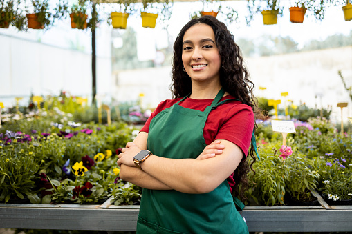 A young, Latina woman works inside a nursery dressed in a green work apron. The dark-skinned woman poses with her arms crossed as she looks at the camera happily. Concept of working woman.