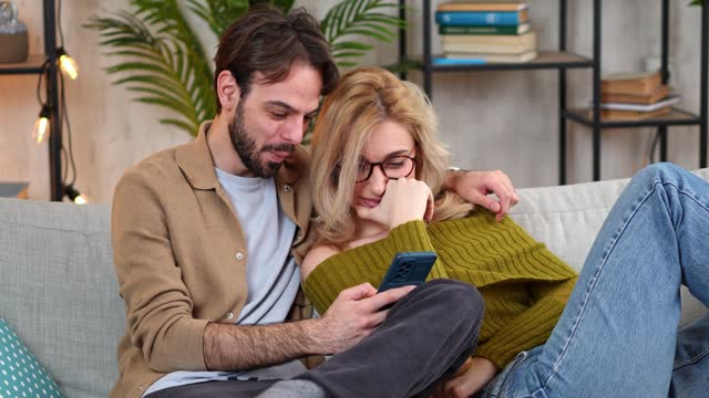 Young Couple Embracing On The Sofa And Using Phone At Home