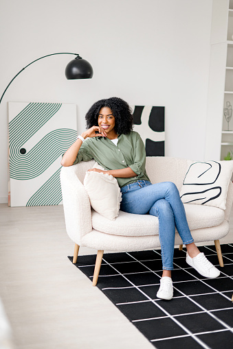 Seated confidently on a minimalist couch, this African-American woman with a radiant smile and relaxed pose provides a perfect representation of modern, casual living in a stylishly decorated space.