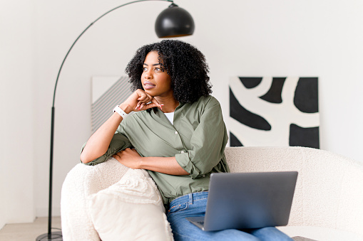 Thoughtful African-American woman sitting on a couch, laptop on her laps, contemplating her next move. The concept highlights the flexibility and contemplation in modern work-from-home scenarios