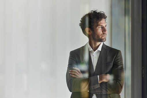 Young male entrepreneur day dreaming while standing with his arms crossed in the office. The view is through glass. Copy space.