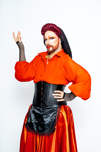 A man in a costume with vibrant red hair and bold makeup, portraying a queer drag queen in a studio setting.