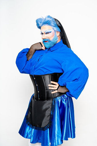 A drag queen poses in a studio wearing a bright blue outfit and matching wig looking at camera