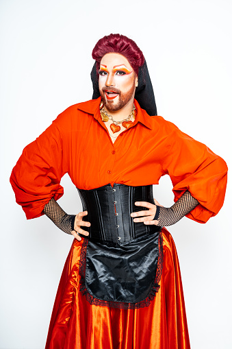 An androgynous drag queen confidently posing in a studio wearing an red shirt and a black skirt.