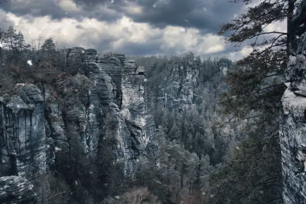 Rugged rocks at Basteibridge during snowfall. Wide view over trees and mountains. National park in Germany