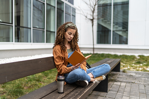 Young university student sitting on bench in the campus and reading a book
