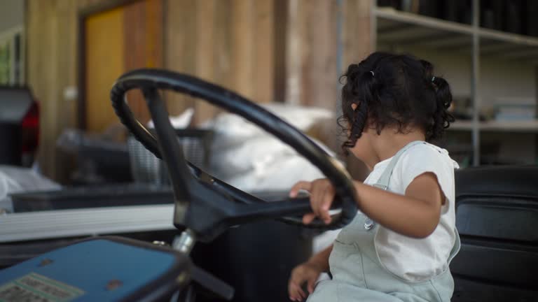 Happy asian children girl playing with lawn mower car, smiling of little cute curly girl having fun activity outdoor, toddler sitting on tractor.