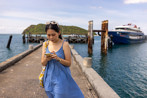Thai woman using smart phone while standing on a pier by the sea outdoors.