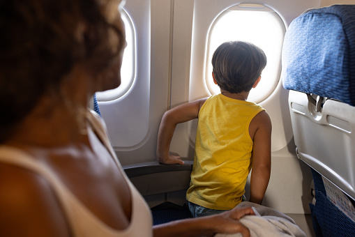 Cute little boy sitting next to his mother during a flight in airplane, he is looking out the window.