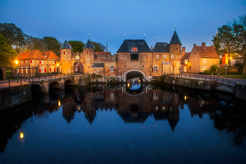 As the sun sets over the historical center of Amersfoort, the iconic Koppelpoort gate and medieval town wall stand tall, overlooking the serene waters of the Eem river. The colorful sky paints a stunning backdrop to this centuries-old landmark, adding to the enchanting atmosphere of the historical center. This image captures the timeless beauty and romantic allure of Amersfoort's medieval architecture against the backdrop of a vibrant sunset.