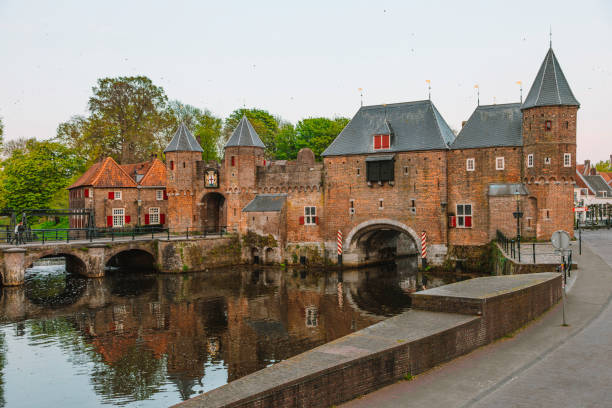 A picturesque view showcases the medieval Koppelpoort gate and town wall alongside the tranquil waters of the Eem river in the historical center of Amersfoort, Netherlands. This iconic landmark, with its imposing architecture and rich history, adds to the timeless allure of Amersfoort's historical center. The image invites viewers to explore the quaint cobblestone streets and preserved medieval architecture that characterize this charming Dutch town.