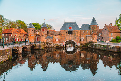 A picturesque view showcases the medieval Koppelpoort gate and town wall alongside the tranquil waters of the Eem river in the historical center of Amersfoort, Netherlands. This iconic landmark, with its imposing architecture and rich history, adds to the timeless allure of Amersfoort's historical center. The image invites viewers to explore the quaint cobblestone streets and preserved medieval architecture that characterize this charming Dutch town.
