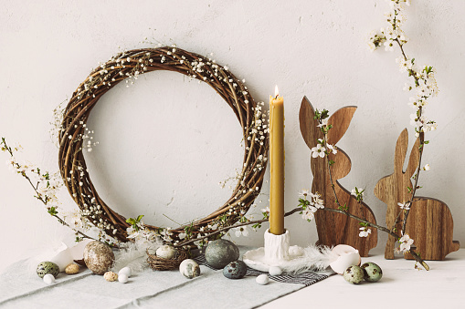 Happy Easter! Stylish easter eggs, wooden bunnies, cherry blossom and candle on rustic table. Modern natural dyed marble eggs and spring flowers. Easter still life decor in countryside home