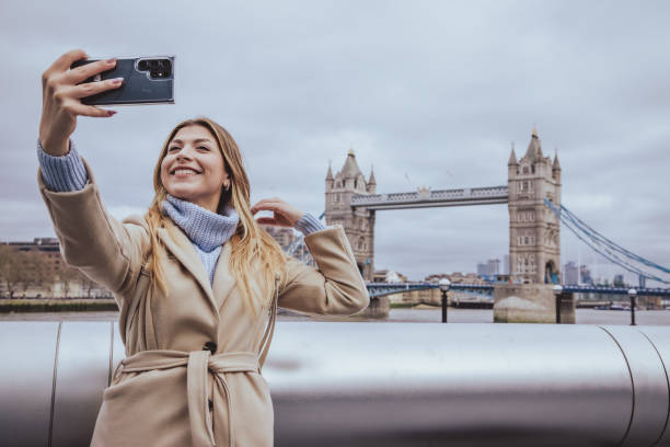 istock Young beutiful blond woman in her 30s smiling while taking selfie with her smart mobile phone device in down town in front of Tower Bridge, City of London, England - creative stock photo 2057879379