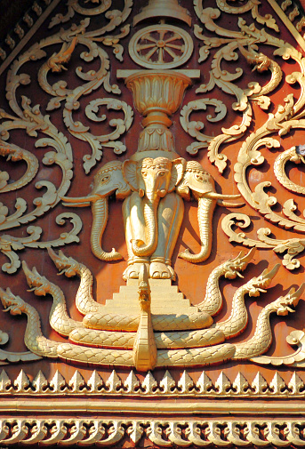 Vientiane, Laos: Wat That Luang Neua, temple on That Luang Park, Pha That Luang complex, Ban Thatluang - ornate golden wood carved gable on a red base. Erawan, the three-headed elephant over the 7 seven headed Naga (Mucalinda, king of Nagas) and under the Dharma wheel, framed by a floral pattern. The Erawan was the central element of the flag of the Kingdom of Laos
