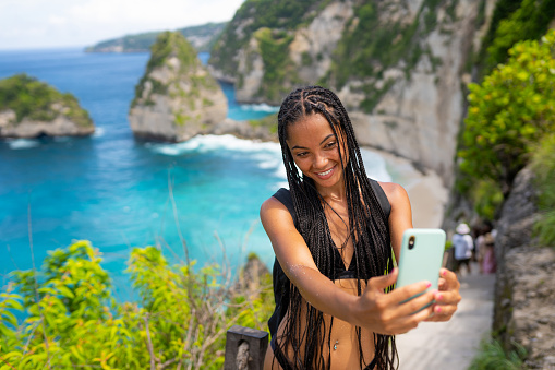 Young asian woman taking selfie or having video call at Kelingking beach. Blues and green sand and the beautiful mountain shaped on the sea.Travel destinations vacations people sharing social media concept nature scenics