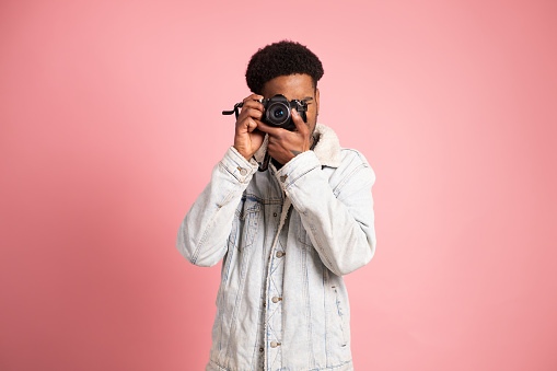 Studio portrait with pink background of a young african man standing using a digital camera