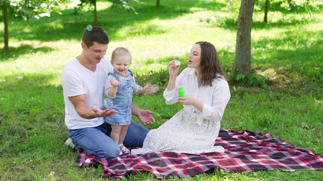 Parents and child have fun, enjoy nature together, sitting on a blanket and blowing bubbles during an outdoor picnic. Cheerful, carefree childhood, family weekends.