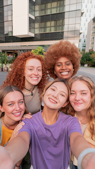 Vertical portrait. A young women group taking a selfie and looking at camera smiling and having fun together outdoor on a sunny day. Multicultural teenage friends. Joyful females laughing outside. High quality photo