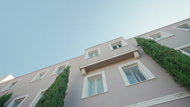 Traditional facade view of Italian residential building with long rectangular windows, without balconies and decorated with high columns of evergreen white cedars against clear blue sky background