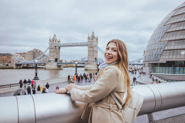 Wide panoramic angle outdoors portrait of a beautiful light haired white woman in her 30s posing on Southbank river Thames with toothy wide smile looking directly in camera and having fun exploring the capital city of London, England. Selective focus technique used with priority on the model and defocused/blurred skyline with recognisable architecture of Tower Bridge and down town and accidental/unrecognizable visitors. Image created during cold season and the model is dressed in warm casual clothes in light cream colours, wearing a leather back pack on a cloudy day during cold season - creative stock photo panoramic wide angle portrait of a young light haired woman smartly dressed in her 30s posing and looking in camera with a wide smile in front of the modern down town skyline, as having fun exploring the capital city of london, uk - creative stock photo