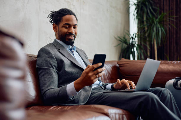 istock Happy black businessman texting on cell phone while working on laptop in hotel lobby. 2057970356