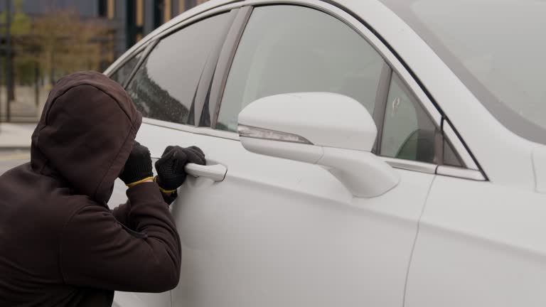 A thief dressed in a black hoodie uses a screwdriver to forcefully pick the lock of a white car door, showcasing a daring act of auto theft. car theft.