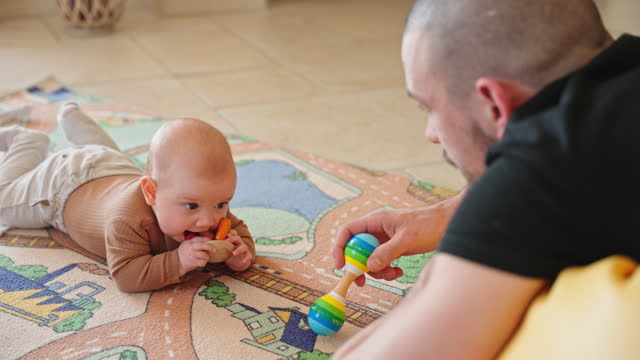 Young Father Shaking Toy Rattle while Playing with Adorable Little Baby Girl at Home