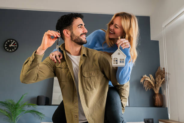 joven matrimonio disfrutando de su nueva casa. está dando un paseo a cuestas a su esposa y están sonriendo y riendo, sosteniendo las llaves de su apartamento, uniéndose y con entusiasmo por nuevos comienzos - jovenes vivienda fotografías e imágenes de stock