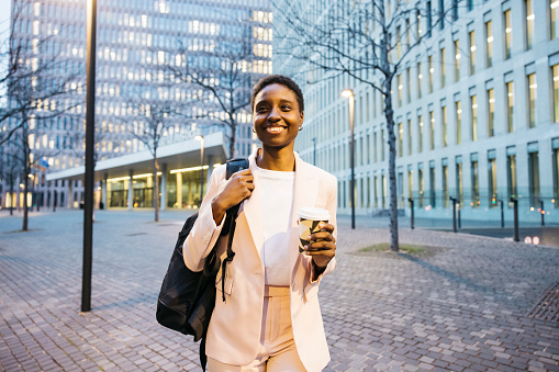 Young beautiful businesswoman with walking on the street with a backpack and a coffee. African businesswoman going to work in the morning.