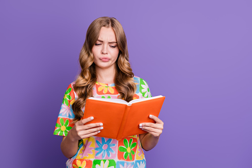 Photo of minded clever student with wavy hair wear print t-shirt thoughtfully look at book in arms isolated on purple color background.