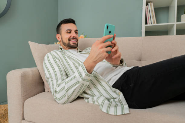 istock Male relax on the sofa in his apartment using his smartphone sending messages. Man lying on the couch and holding the phone using and application and surfing online in the living room 2058140123