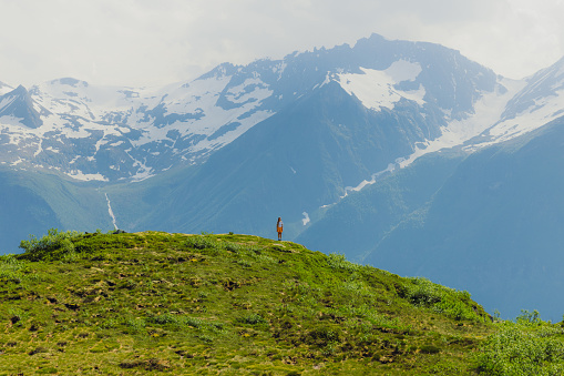 Distant view of female backpacker in orange shorts staying at the edge of the green mountain admiring dramatic view of blue peaks in the background in Sunnmore Alps, Western Norway