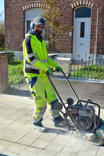Leuven, Flemish-Brabant, Belgium- March 4, 2024:finished the road works. Worker in yellow reflective jacket and pants pushing a manual concrete paving leveling machine that is damming stone pavement